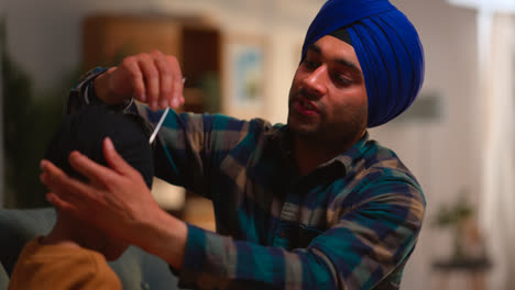 Father-Using-Salai-Needle-To-Tidy-Away-Hair-When-Tying-Turban-Onto-Head-Of-Young-Sikh-Son-Sitting-On-Sofa-Shot-in-Real-Time